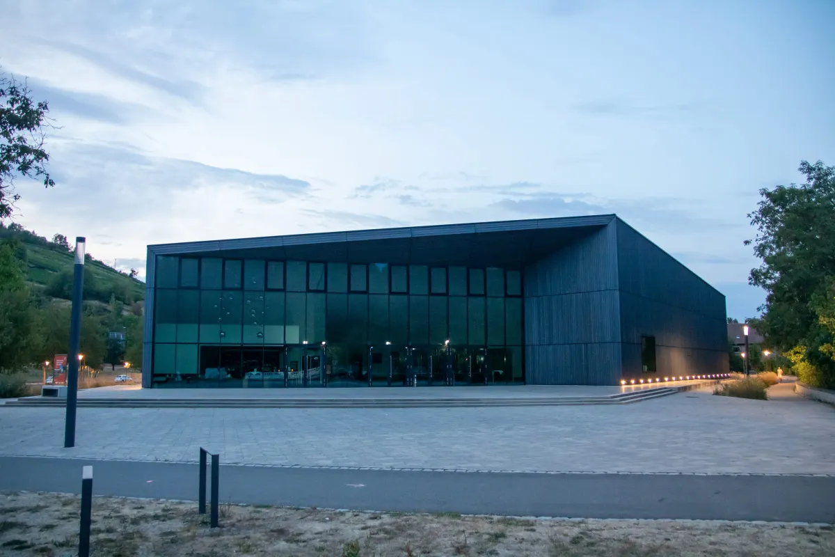 Die TauberPhilharmonie in der blauen Abenddämmerung, ein moderner Bau mit dunkler Holzverschalung und großer Glasfront. Das erleuchtete Foyer scheint nach außen, entlang der Vorplatzkante leuchtet eine Reihe kleiner Bodenlichter. Links ein begrünter Weinberg, davor ein weiter gepflasterter Platz.