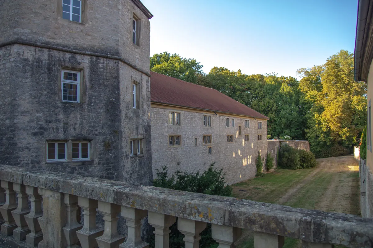 Seitenansicht des Schlosses Weikersheim von einer steinernen Balustrade aus. Links ein massiver Natursteinbau mit Sprossenfenstern, daneben ein langgestreckter Flügel mit rotem Ziegeldach. Rechts eine Wiese und ein von Bäumen gesäumter Weg im Abendlicht.