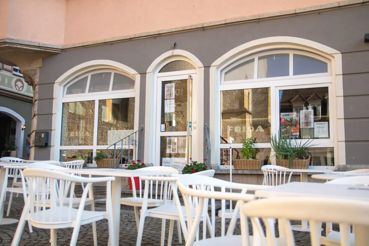 Blick auf die graue Ladenfront des FRIDA café und deli mit zwei großen Rundbogenfenstern und der schmalen Glastür in der Mitte. Auf den Scheiben der Schriftzug FRIDA, im Fenster Plakate und Aushänge. Davor leere weiße Stühle und Tische auf Kopfsteinpflaster, an der Treppe rote Geranien.