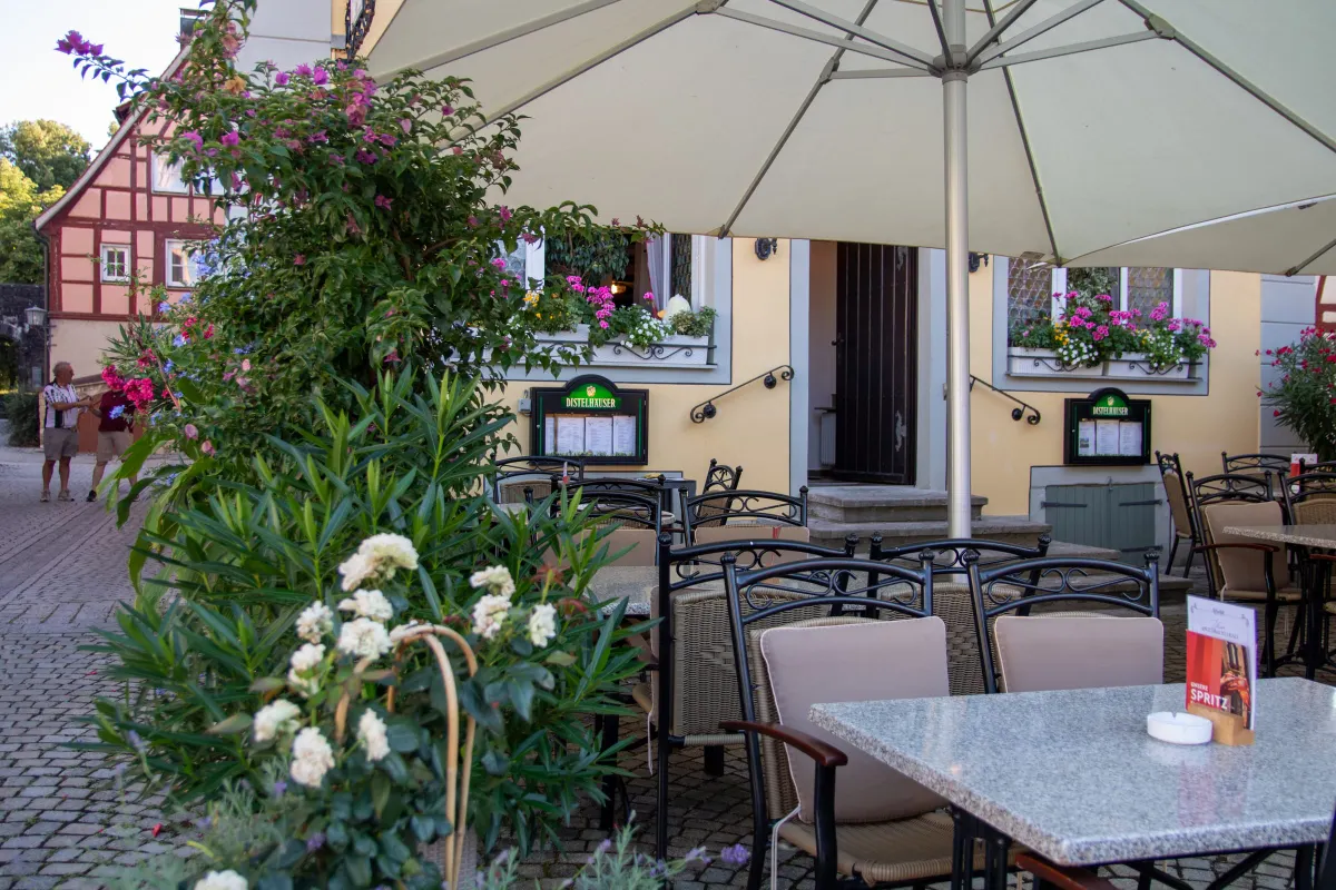 Terrasse mit Korbstühlen und Steintischen unter einem Sonnenschirm am Abend. Im Hintergrund die gelbe Fassade mit Blumenkästen und Distelhäuser Schaukästen, links ein rotes Fachwerkhaus und zwei stehende Passanten.