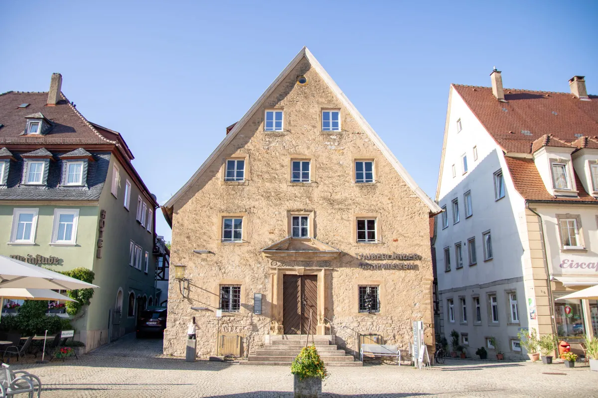 Hoher historischer Natursteinbau des Tauberländer Dorfmuseums mit spitzem Giebel, kleinen Sprossenfenstern und einem steinernen Portal mit Holztür, zu dem eine Treppe führt. An der Fassade der Schriftzug Tauberländer Dorfmuseum, flankiert von Nachbarhäusern am gepflasterten Marktplatz.