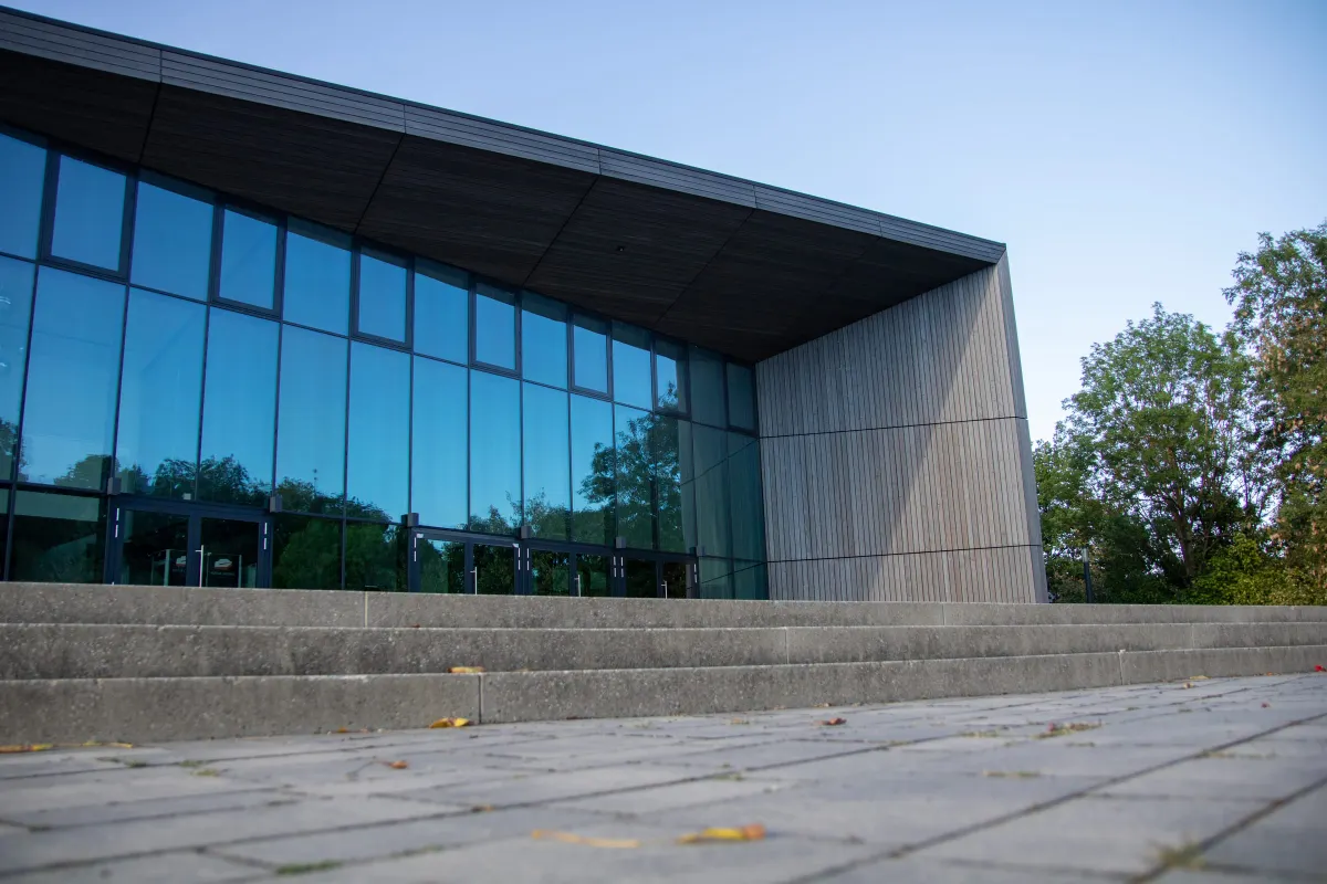 Seitliche Perspektive der TauberPhilharmonie aus tiefer Sicht vom gepflasterten Vorplatz. Die große blau spiegelnde Glasfassade mit dunklen Rahmen und das weit auskragende Holzdach verlaufen schräg ins Bild, rechts steht ein Baum vor blauem Himmel.