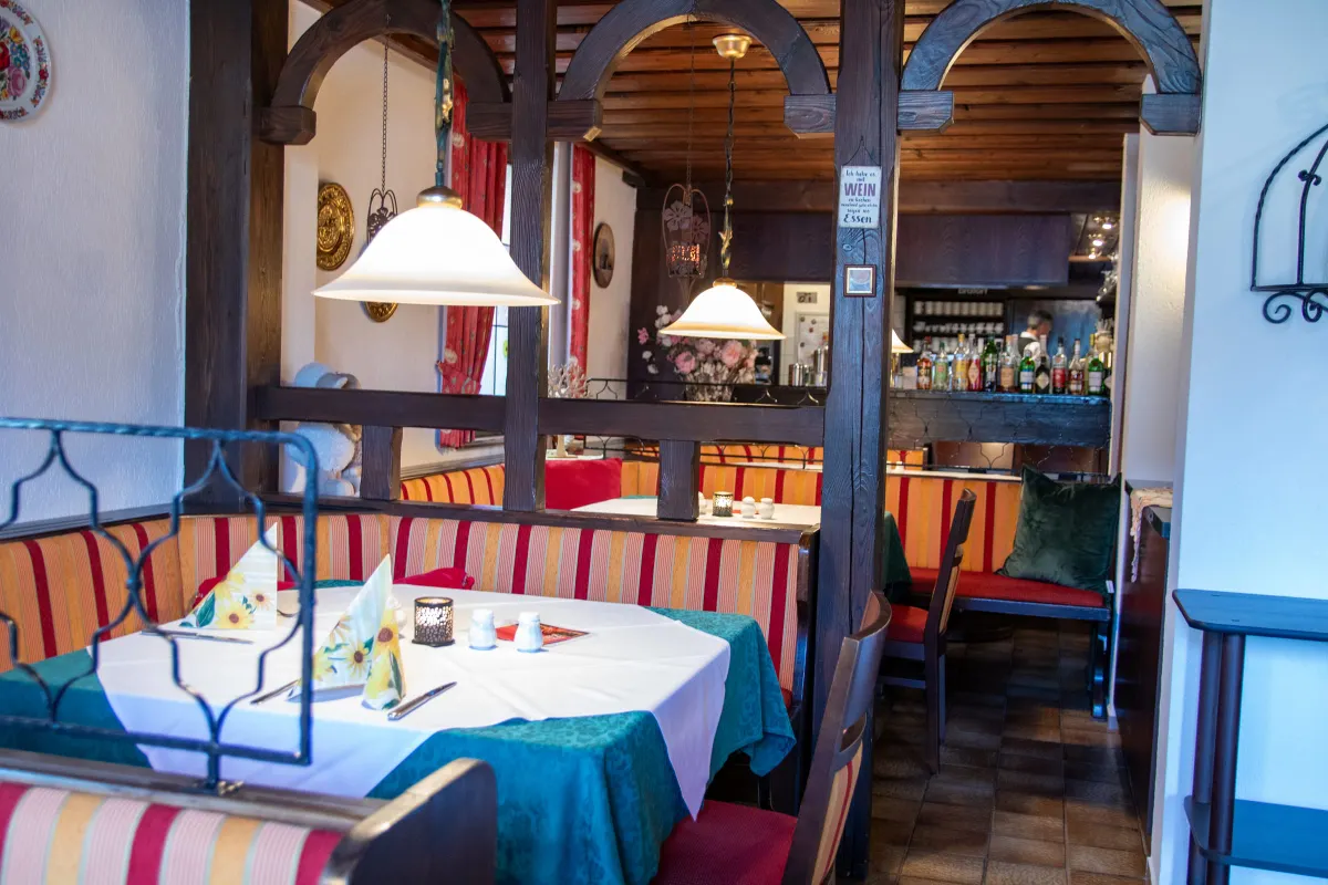 Gemütliche Gaststube mit dunklen Holzbögen und Fachwerkbalken. Ein eingedeckter Tisch mit weißer und grüner Decke steht an gestreiften Sitzbänken. Im Hintergrund eine Bar mit Flaschen und eine Hängelampe mit hellem Glasschirm.