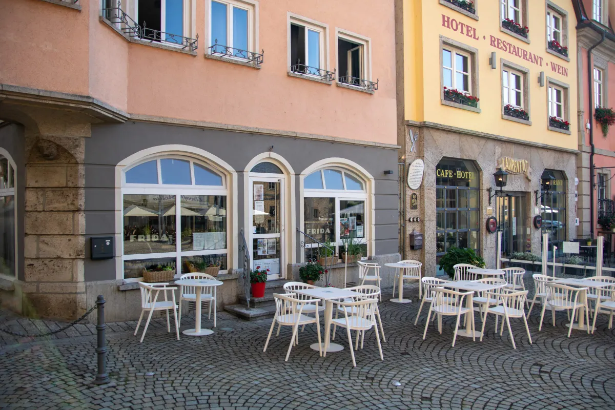 Terrasse des FRIDA café und deli am gepflasterten Marktplatz mit weißen Bistrotischen und Stühlen. Die graue Ladenfront hat drei rundbogige Fenster mit dem Schriftzug FRIDA, davor Kräuterkörbe und ein roter Geranientopf. Rechts eine gelbe Hotelfassade mit der Aufschrift Hotel Restaurant Wein.