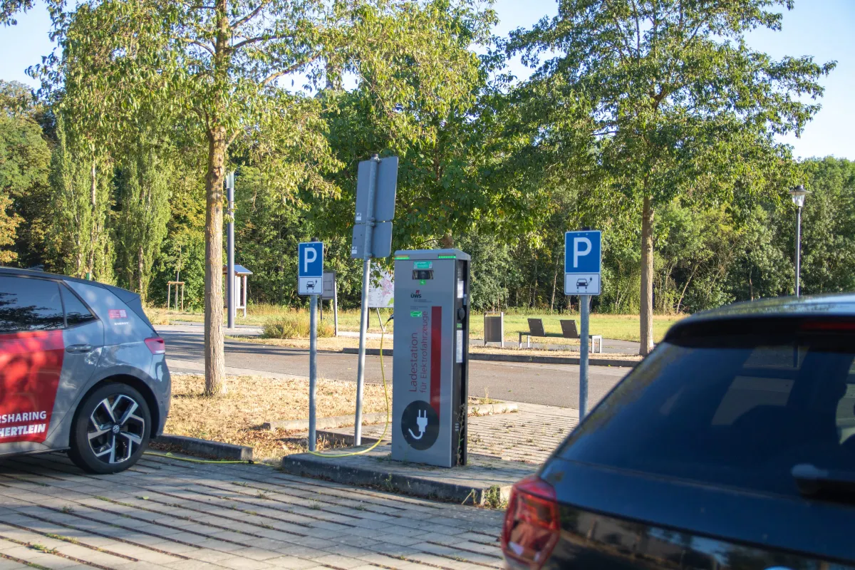 Ladestation für Elektrofahrzeuge auf dem Parkplatz Tauberwiesen, eine graue Säule mit rotem Streifen und Steckersymbol. Daneben zwei blaue Parkschilder mit E-Auto-Symbol. Links ein grau-rotes Carsharing-Fahrzeug am Ladekabel, rechts ein dunkles Auto. Im Hintergrund Bäume und eine Bank.
