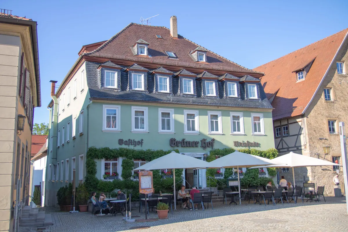 Grün gestrichenes Gasthaus mit hohem Mansarddach und vielen Dachgauben, an der Fassade in schwarzer Schrift Gasthof, Grüner Hof und Weinstube sowie berankte Fenster. Davor auf dem gepflasterten Platz eine Terrasse mit weißen Sonnenschirmen, Tischen und Gästen.