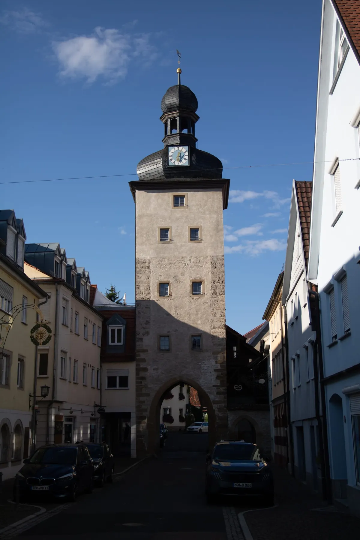 Hoher steinerner Torturm mit rauem Bruchsteinmauerwerk und wenigen kleinen Fenstern, oben eine schwarze Zwiebelhaube mit Laterne und Wetterfahne sowie eine Turmuhr mit blauem Zifferblatt. Unten ein rundbogiges Durchfahrtstor, das eine schmale Gasse mit parkenden Autos rahmt.