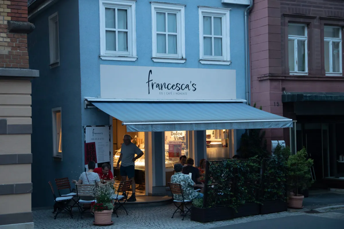 Blaue Fassade des Eiscafés francesca's am Abend mit dem geschwungenen Schriftzug francesca's, Eis, Café, Momente und einer gestreiften Markise. Unter der Markise sitzen Gäste an Tischen, im hell erleuchteten Eingang ist die Eistheke zu sehen, rechts ein rosa Nachbarhaus mit der Hausnummer 15.