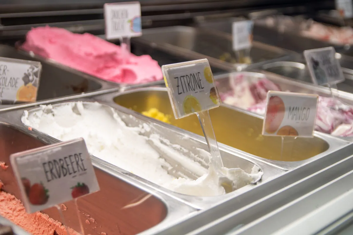 Blick in die Eistheke des francesca's mit mehreren Sorten in Edelstahlbehältern, darunter Erdbeere, Zitrone und Mango. Kleine beschriftete Schilder mit Fruchtbildern stecken im Eis, im Hintergrund eine pinke und eine weiße Sorte.