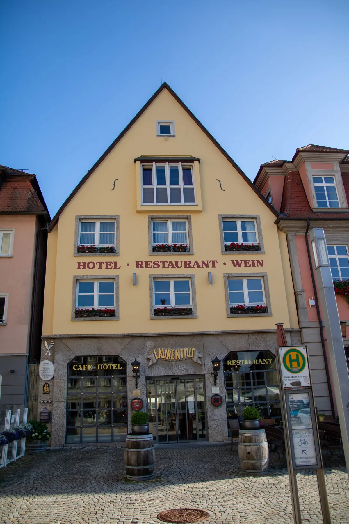 Der hohe spitze Giebel des Hotels Laurentius vor blauem Himmel, gelbe Fassade mit rotem Schriftzug HOTEL, RESTAURANT, WEIN und roten Geranien an den Fenstern. Unten der Eingang mit goldenem Laurentius-Schild, davor Holzfässer und ein grünes Haltestellenschild auf dem gepflasterten Marktplatz.