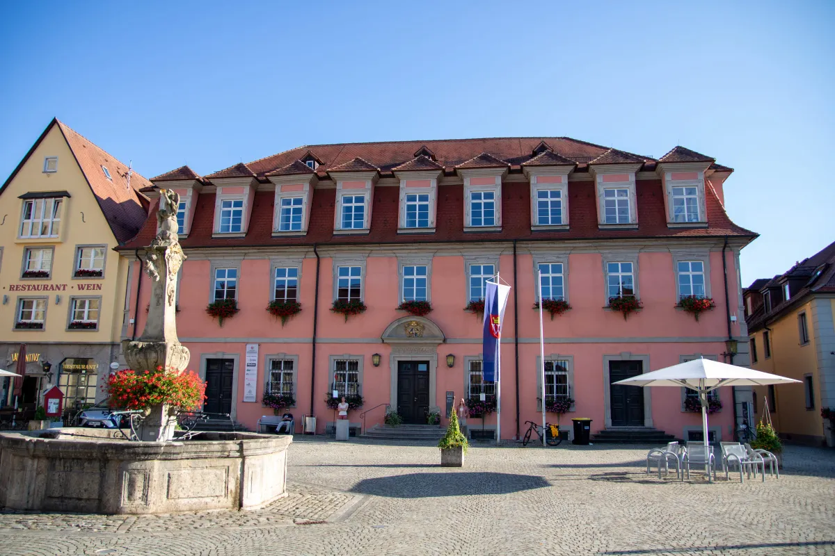 Langgestrecktes barockes Rathaus mit rosa verputzter Fassade, rotem Mansarddach mit Gauben und rot blühenden Blumenkästen an den Fenstern. Über dem Eingang ein Wappengiebel, davor eine blaue Fahne. Auf dem gepflasterten Platz stehen ein Steinbrunnen mit Figur und ein weißer Sonnenschirm.