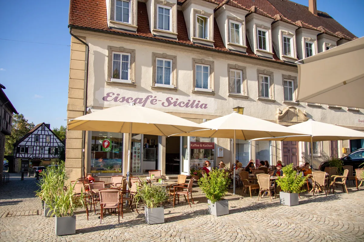 Historisches Gebäude mit heller Fassade und rotem Mansarddach, an der Wand der geschwungene Schriftzug Eiscafé-Sicilia. Davor eine Terrasse mit großen cremefarbenen Sonnenschirmen, Korbstühlen, Tischen und Gästen auf gepflastertem Boden, ein Schild wirbt für Kaffee und Kuchen.