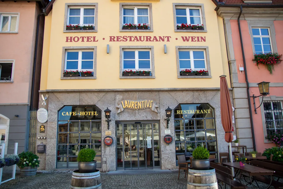 Gelbe Fassade des Café-Hotel-Restaurants Laurentius am Marktplatz, mit dem Schriftzug HOTEL, RESTAURANT, WEIN und roten Geranien in den Fensterkästen. Über dem verglasten Eingang das goldene Schild LAURENTIVS, davor eine gepflasterte Terrasse mit Holzfässern, Kübelpflanzen und Tischen.