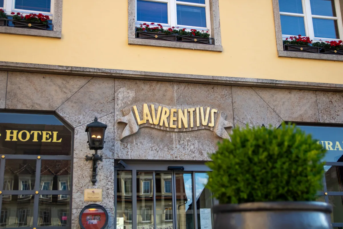 Nahaufnahme des goldenen Schriftzugs LAURENTIVS auf einem Steinband über dem Eingang, flankiert von einer schmiedeeisernen Wandlaterne. Links und rechts die goldenen Wörter HOTEL und RESTAURANT auf den Schaufenstern, oben rote Geranien in den Fensterkästen.