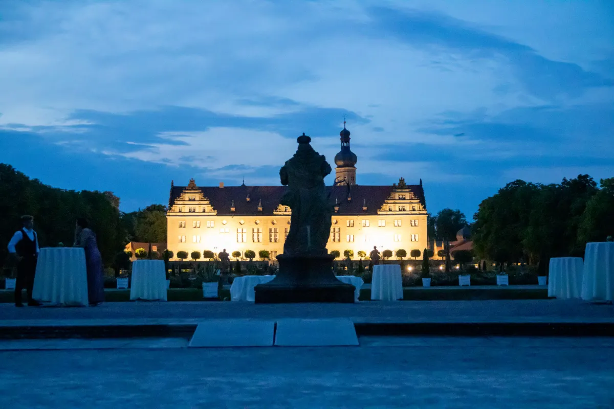 Das Schloss Weikersheim in der blauen Abenddämmerung, die lange Fassade und der Turm mit Zwiebelhaube warm angestrahlt. Im Schlossgarten stehen weiß eingedeckte Stehtische für eine Abendveranstaltung, im Vordergrund die dunkle Silhouette einer Gartenfigur.