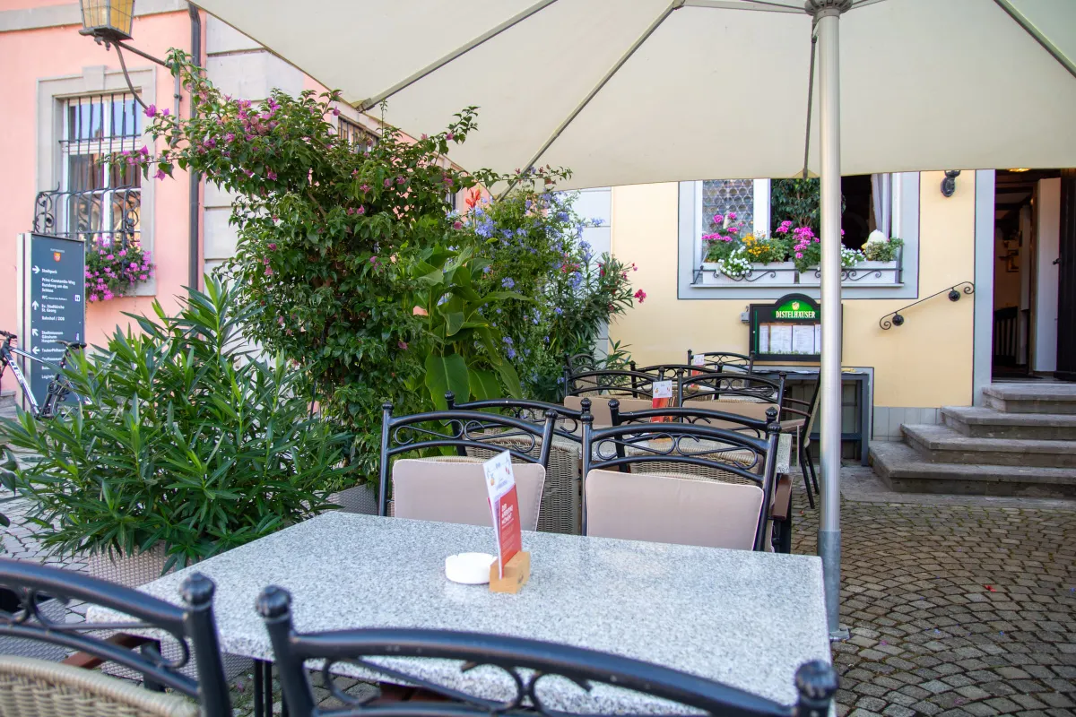 Terrasse unter einem großen Sonnenschirm mit Korbstühlen und einem Steintisch auf Kopfsteinpflaster. Links blühende Kübelpflanzen, im Hintergrund die gelbe Hauswand mit Blumenkästen, ein Distelhäuser Schaukasten und ein Wegweiser.