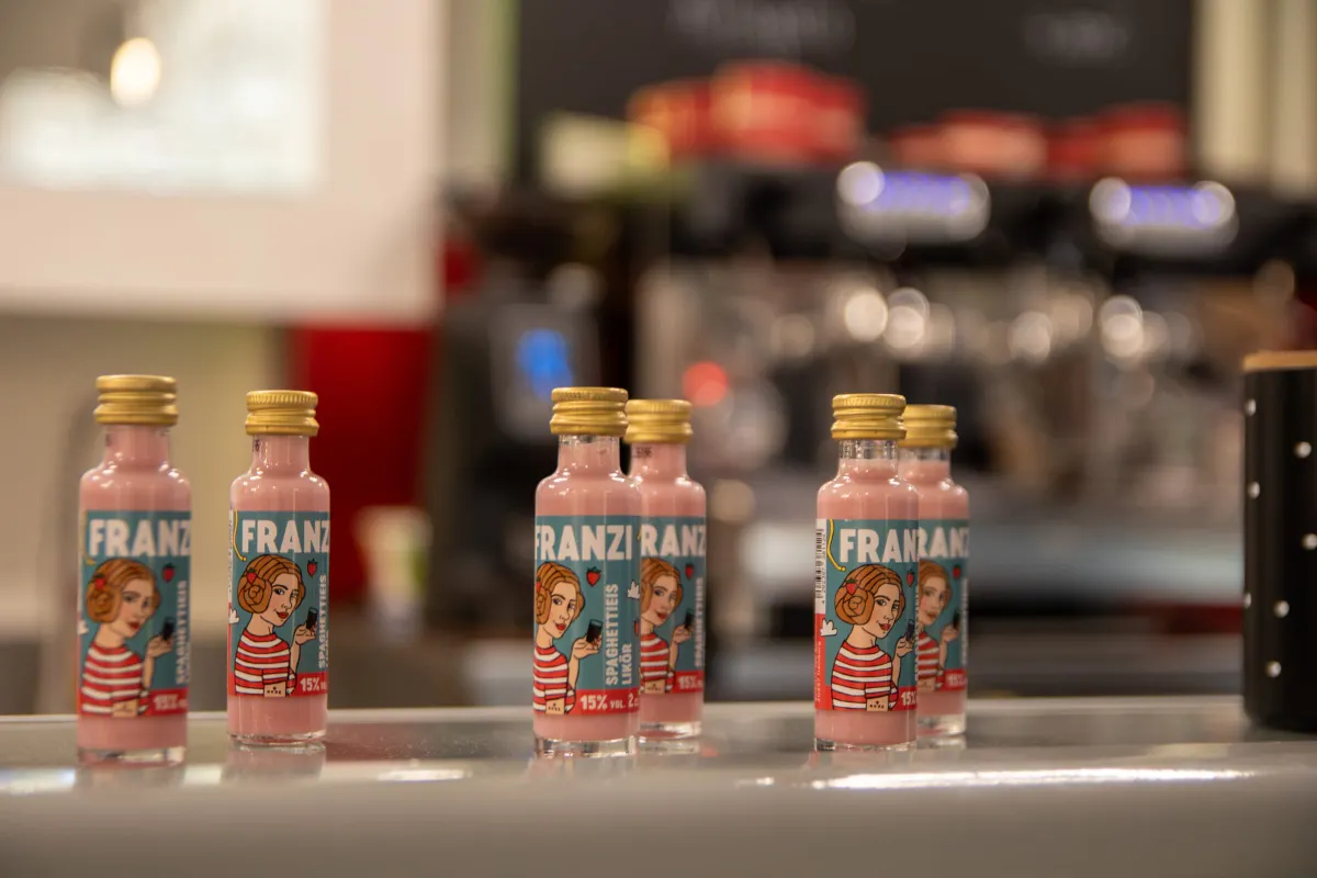 Nahaufnahme mehrerer kleiner Flaschen mit rosa Likör auf einer Theke im francesca's. Die türkisen Etiketten zeigen eine gezeichnete Frau im rot-weiß gestreiften Shirt und den Schriftzug FRANZI, Spaghettieis-Likör, 15% vol. Im unscharfen Hintergrund eine Siebträger-Espressomaschine.