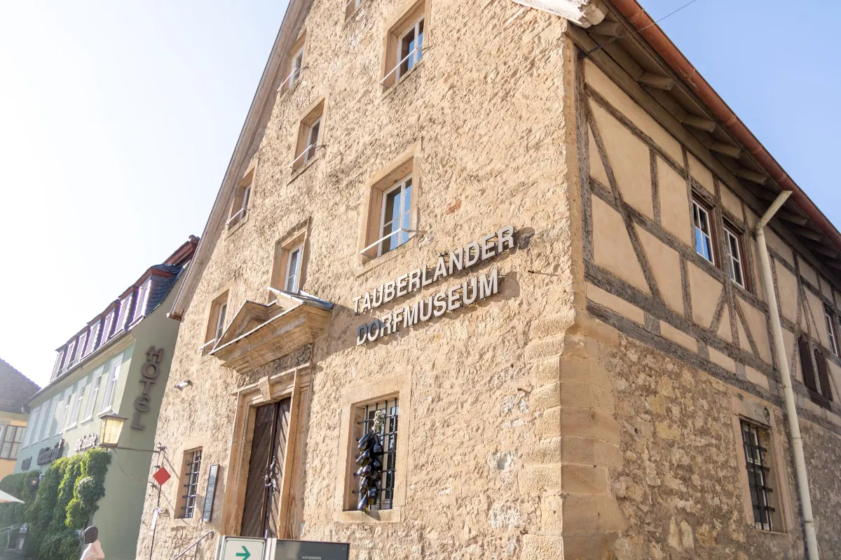 Schräge Eckansicht des Tauberländer Dorfmuseums von unten. Links die verputzte Natursteinfassade mit vergitterten Fenstern und dem Schriftzug Tauberländer Dorfmuseum, rechts eine Fachwerkwand mit dunklen Balken unter blauem Himmel. Links im Hintergrund ein Nachbargebäude mit Hotel-Schild.