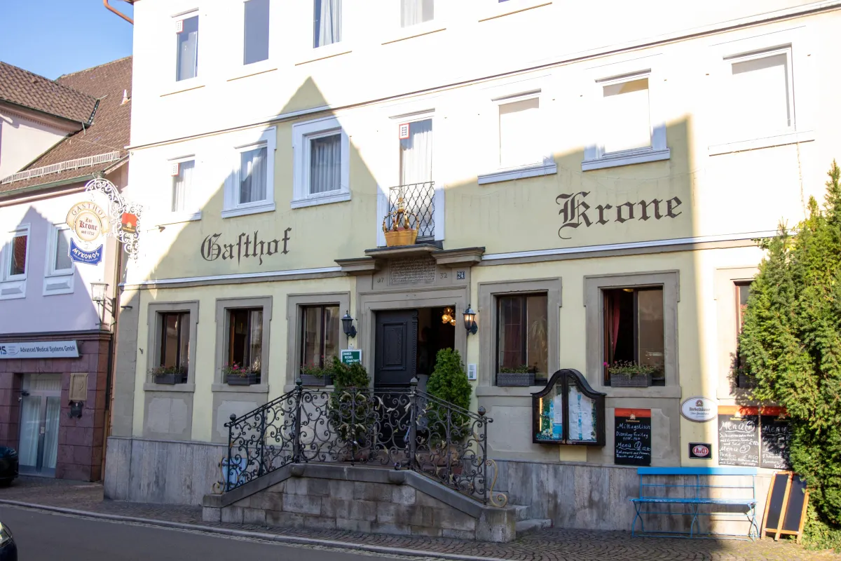 Gelb gestrichene Fassade des Gasthofs zur Krone mit den aufgemalten Wörtern Gasthof und Krone in dunkler Schrift. Über dem Eingang eine goldene Krone als Wappen. Eine geschwungene schmiedeeiserne Treppe führt zur dunklen Haustür, daneben ein hängendes Wirtshausschild mit dem Zusatz Mykonos.