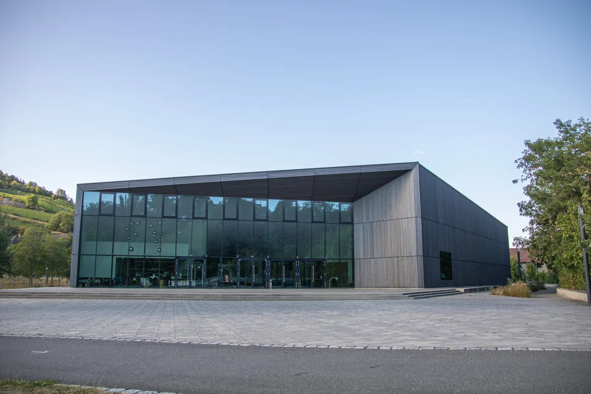 Moderner Konzertsaalbau der TauberPhilharmonie in Weikersheim mit langgestreckter Fassade aus dunkler Holzverschalung und einer hohen Glasfront zum gepflasterten Vorplatz. Links im Hintergrund ein begrünter Weinberg, rechts ein Baum unter blauem Himmel.