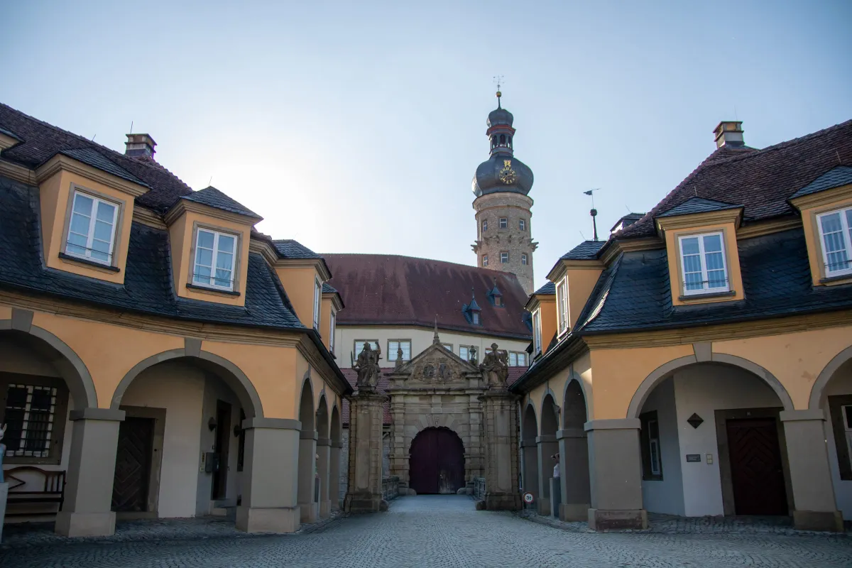 Blick durch den gepflasterten Vorhof des Schlosses Weikersheim mit den beiden gelb verputzten Arkadengebäuden links und rechts. In der Mitte das steinerne Torhaus mit Rundbogenportal, flankiert von zwei Figuren. Dahinter ragt der Schlossturm mit welscher Zwiebelhaube und Turmuhr in den Himmel.