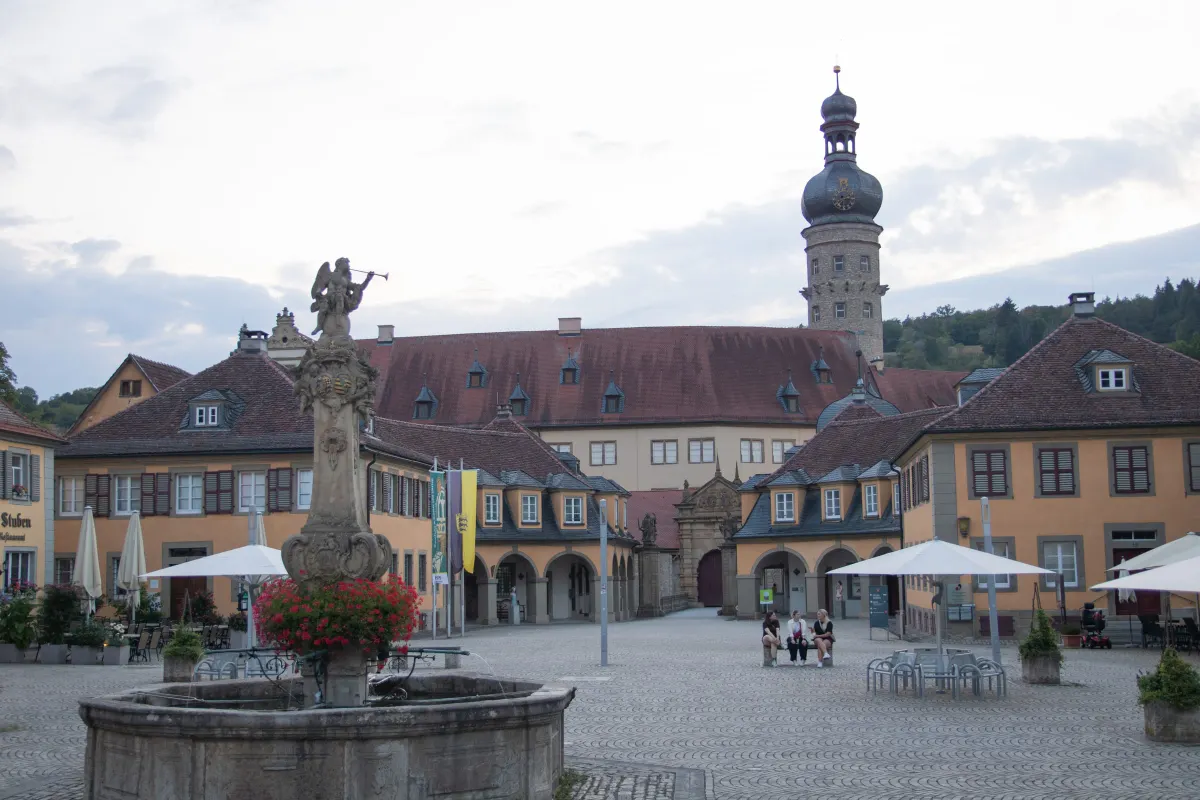 Der Marktplatz vor dem Schloss Weikersheim mit einem steinernen Brunnen, dessen Säule eine Engelsfigur und rote Geranien trägt. Dahinter die gelben Arkadengebäude und das Torhaus, rechts erhebt sich der Schlossturm mit Zwiebelhaube vor bewölktem Himmel, links ein Restaurant mit Sonnenschirmen.