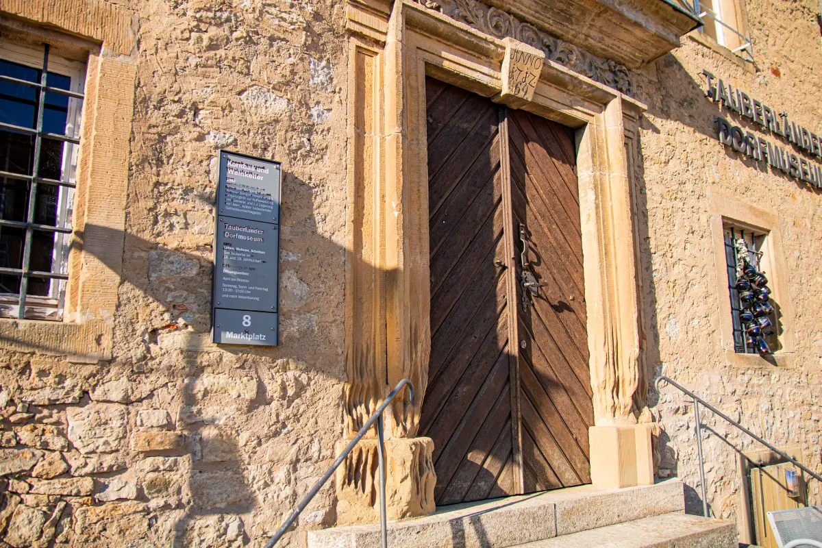 Steinernes Rundbogenportal des Tauberländer Dorfmuseums mit hoher Holztür aus diagonalen Brettern und geschnitztem Türsturz mit Jahreszahl. Links an der Natursteinfassade eine graue Info-Tafel zu Kornbau und Weinkeller, rechts ein vergittertes Fenster, davor Steinstufen mit Geländer.