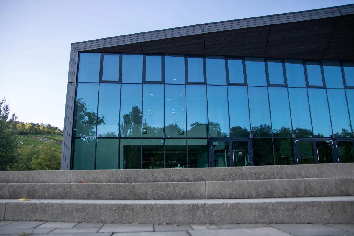Nahansicht der Glasfront der TauberPhilharmonie mit dunklen Holzrahmen und auskragendem Vordach aus Holz. In den Scheiben spiegeln sich Bäume und der Weinberg, dahinter sind hängende Kugelleuchten und das Foyer erkennbar. Davor breite Steinstufen.