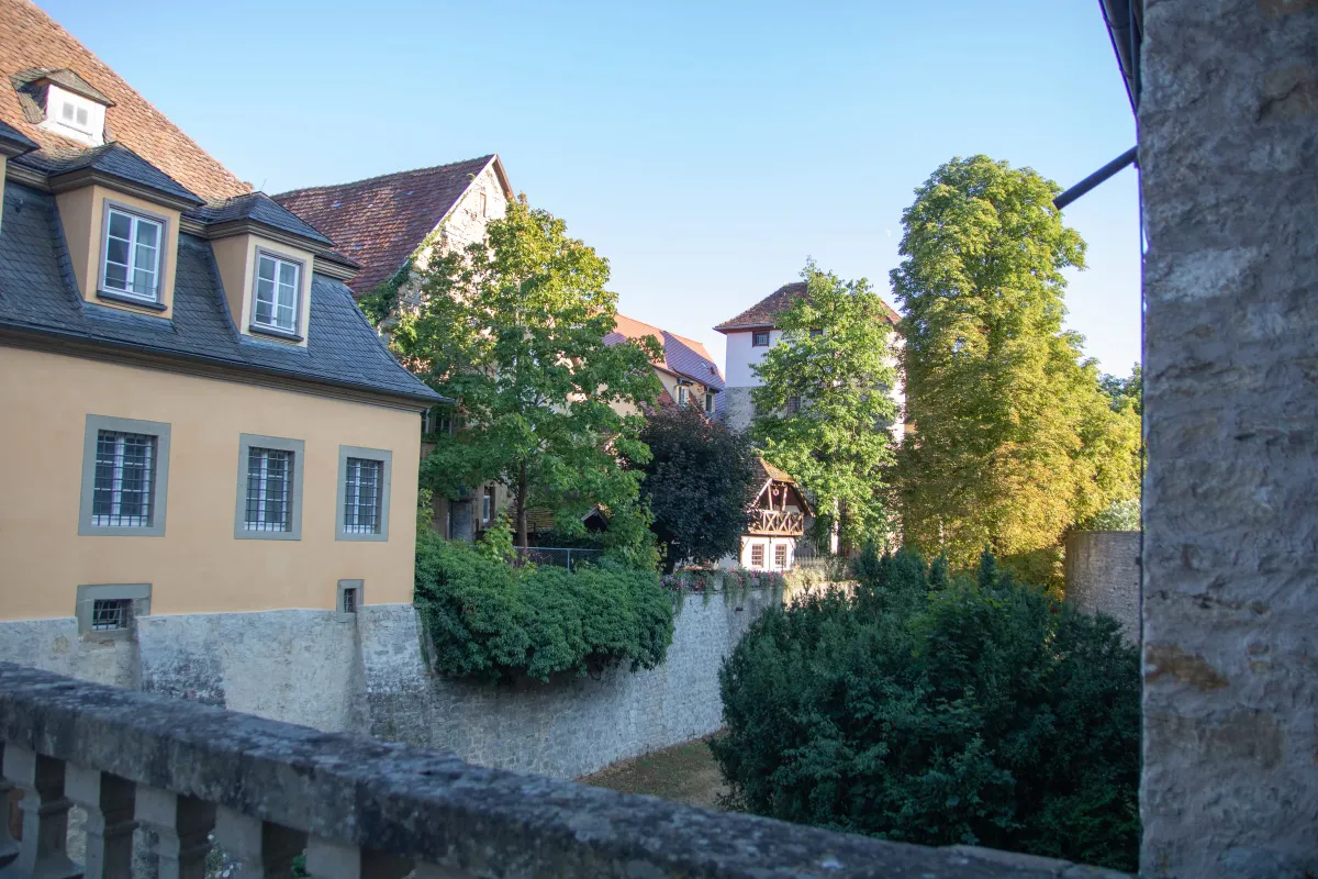 Blick von einer steinernen Balustrade des Schlosses Weikersheim über den Schlossgraben. Links ein gelber Putzbau mit Schleppgauben, dahinter Natursteingebäude und ein kleines Fachwerkhaus zwischen dichten grünen Bäumen und Sträuchern im warmen Abendlicht.