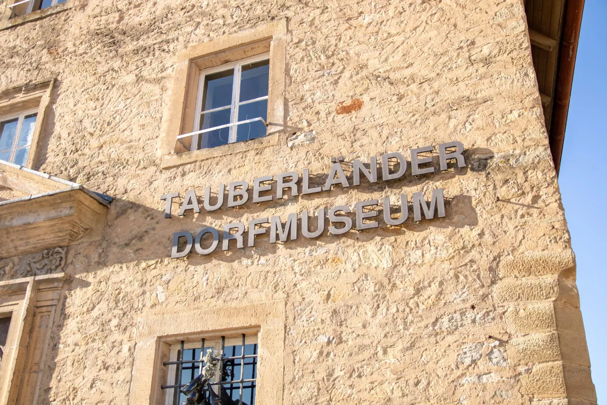 Detailaufnahme der rauen, hellen Natursteinfassade des Tauberländer Dorfmuseums mit dem in Metallbuchstaben angebrachten Schriftzug Tauberländer Dorfmuseum. Darüber ein Sprossenfenster mit Holzrahmen, unten ein vergittertes Fenster, alles im warmen Sonnenlicht.