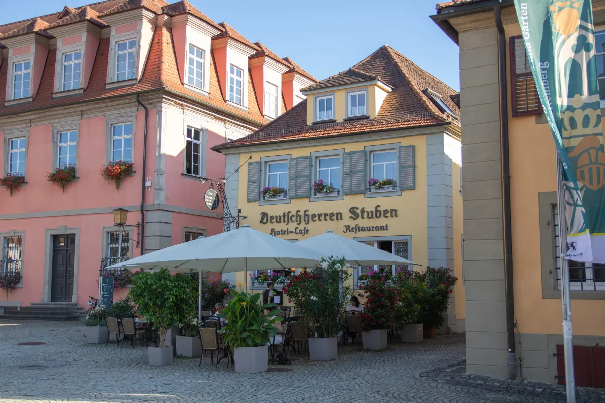 Gelbe Fassade der Deutschherren Stuben mit der Aufschrift Hotel-Cafe Restaurant an einem Platz mit Kopfsteinpflaster. Davor eine Terrasse mit weißen Sonnenschirmen, Tischen und blühenden Kübelpflanzen. Links ein rosafarbenes Gebäude mit roten Geranien.