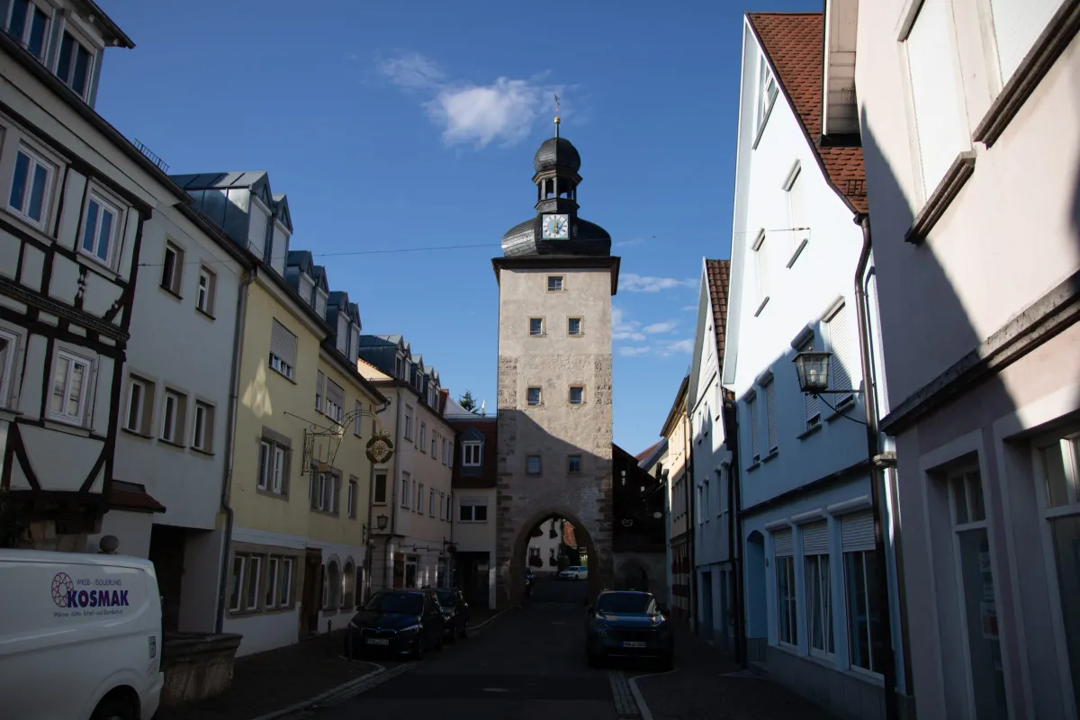 Blick durch eine schmale Altstadtgasse auf den Gänsturm, einen hohen Steinturm mit schwarzer Zwiebelhaube, Laterne und Turmuhr. Links und rechts stehen verputzte Wohn- und Fachwerkhäuser, im Vordergrund parken Autos und ein weißer Transporter am Straßenrand.