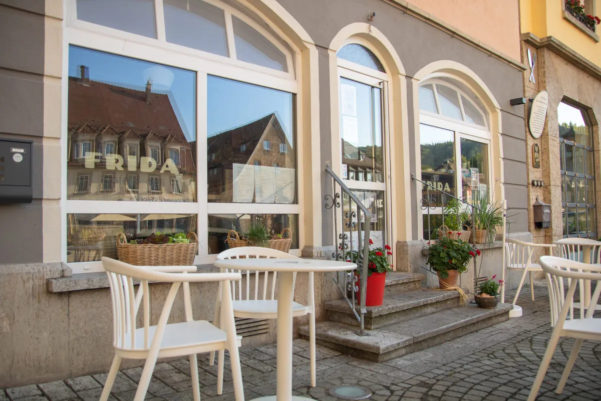Nahansicht der Ladenfront des FRIDA café und deli mit großem Rundbogenfenster und dem Schriftzug FRIDA café und deli auf der Scheibe. Im Glas spiegeln sich Fachwerkhäuser des Marktplatzes. Davor Weidenkörbe mit Kräutern, weiße Stühle und ein roter Geranientopf an der Steintreppe zum Eingang.